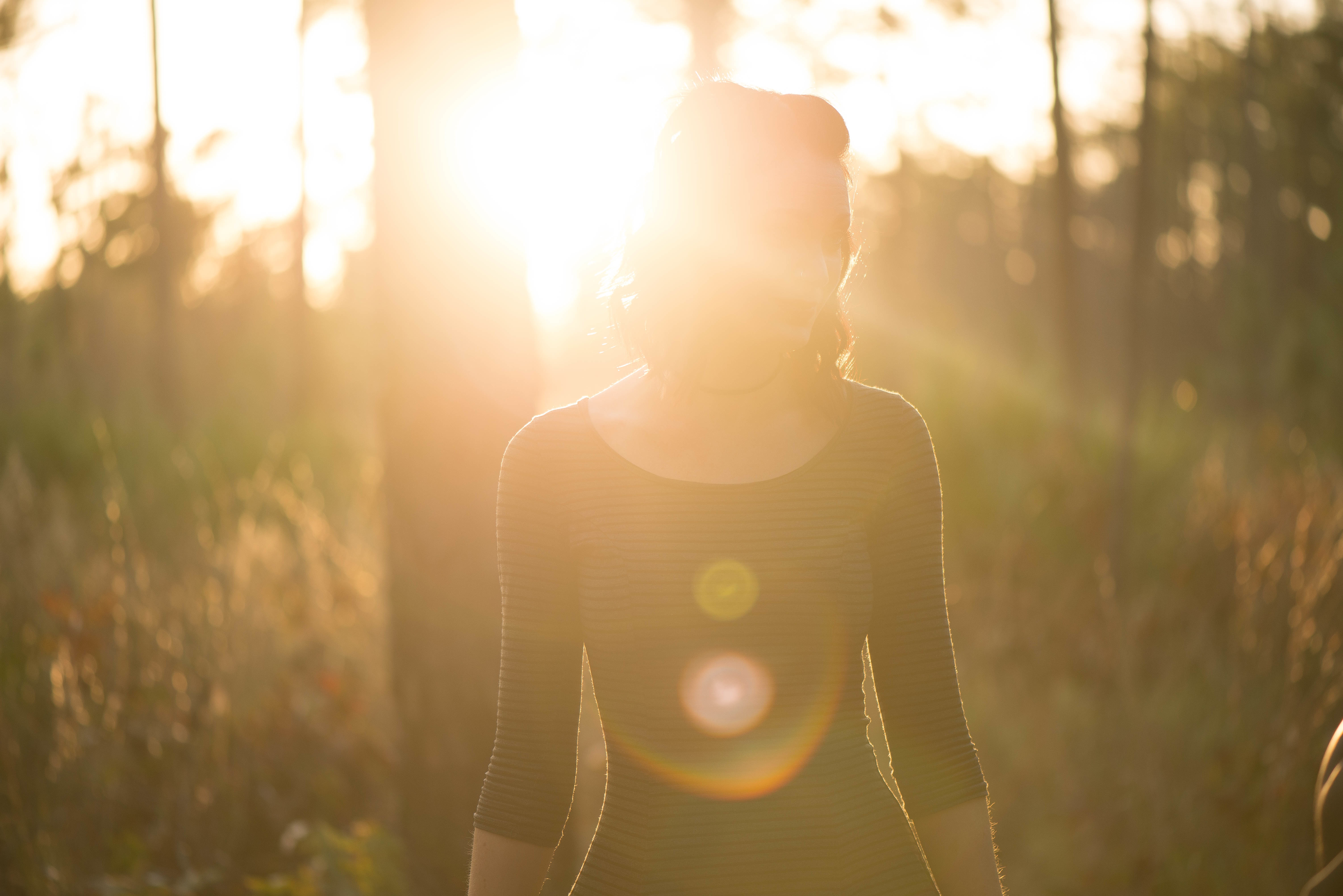 A woman standing in a forest silhouetted by the glare of a setting sun 